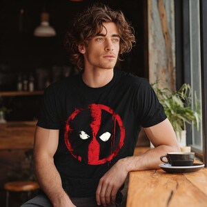 May include: Black t-shirt featuring a red and black graphic of a character's face. The design is a stylized, circular emblem with white eye shapes. The shirt is short-sleeved and made of a soft material.