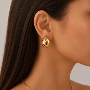 May include: A pair of gold teardrop-shaped earrings. The earrings are smooth, shiny, and reflect light. The earrings are worn on the ear of a person with dark hair. The earrings are part of a jewelry set.