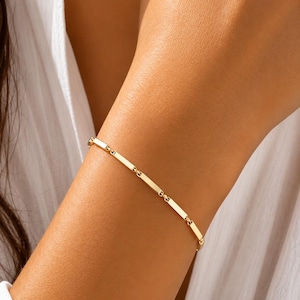 Könnte beinhalten: Ein goldenes Armband mit rechteckigen Gliedern, die durch kleine kreisförmige Glieder verbunden sind. Das Armband wird an einem Arm getragen, mit einem weißen Hemd im Hintergrund. Es ist ein filigranes Schmuckstück.