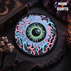 May include: A circular embroidered patch featuring a stylised eye design with a black pupil, a bright green iris, and a light blue sclera. The surrounding area is filled with intricate, swirling patterns in shades of pink, blue, and purple.