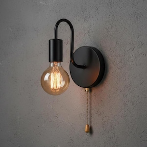 May include: Black wall light with a curved arm and a round, exposed bulb. The fixture has a pull chain with a wooden toggle. The bulb emits a warm amber glow, and the light is mounted on a grey wall.