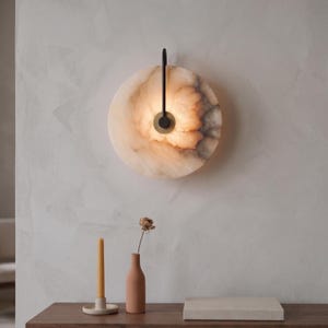 Può includere: Una lampada da parete circolare con un disco in pietra traslucida marmorizzata e illuminazione interna calda. Un sottile braccio in metallo nero opaco si estende dall'alto e si collega a un supporto circolare in ottone al centro.