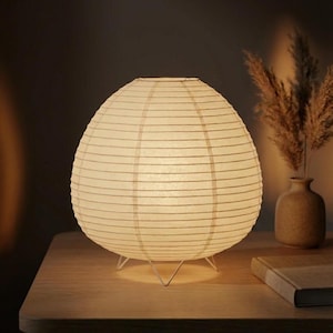 Könnte beinhalten: Cremefarbene Papierlaterne mit kugelförmiger Form, getragen von einem weißen Metallfuß. Die Lampe ist beleuchtet und strahlt ein weiches Licht aus. Sie steht auf einem Holztisch, im Hintergrund eine Vase und ein Buch. Durchmesser ca. 30 cm.