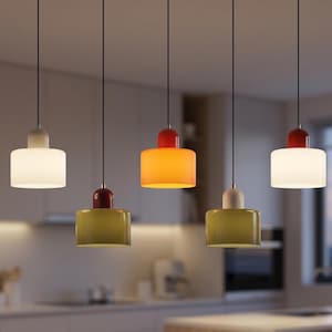 Op de afbeelding: Vijf hanglampen met cilindrische kappen in verschillende kleuren: wit, oranje en groen. Elke lamp heeft een rode of witte bovenkant en hangt aan een zwart snoer. De lampen hangen in een keuken.
