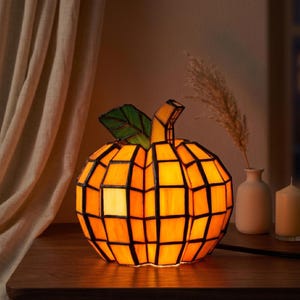 Może przedstawiać: Lampa w kształcie dyni ze szkła witrażowego, emitująca ciepłą pomarańczową poświatę. Lampa ma geometryczny wzór z czarnymi liniami i zielonym liściem. Stoi na drewnianej powierzchni, z neutralnym tłem.
