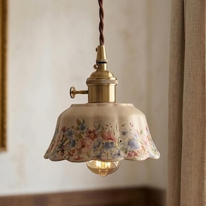 Könnte beinhalten: Vintage-Pendelleuchte mit cremefarbenem, gewelltem Lampenschirm, verziert mit einem Blumenmuster in Rosa, Blau und Lila. Die Leuchte hat eine Messingoberfläche und ein braunes Kabel.