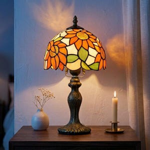 May include: A stained glass table lamp with a sunflower design in orange, green, and white. The lamp has a bronze-colored base and sits on a wooden table. A small white vase with dried flowers and a lit candle are also on the table.