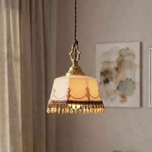 Puede incluir: Lámpara colgante de estilo vintage con pantalla crema con diseño floral marrón. La pantalla está adornada con encaje marrón oscuro y cuentas de cristal. La lámpara tiene un marco de metal dorado y un cable marrón.