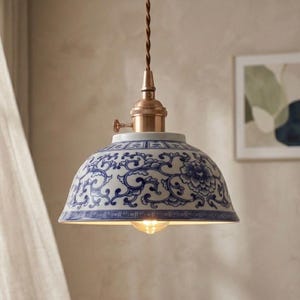 May include: A ceramic pendant light with a blue and white floral design. The shade has a domed shape and is suspended by a brown cable. The top of the fixture is copper-toned. The light bulb is visible. The design is reminiscent of vintage decor.
