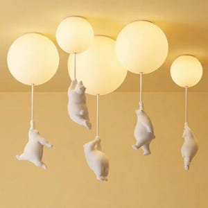 May include: A ceiling light fixture featuring five spherical, off-white lamps. Each lamp is suspended by a thin white rod, with a white bear figurine clinging to the rod below each lamp. The bears appear to be climbing or hanging.