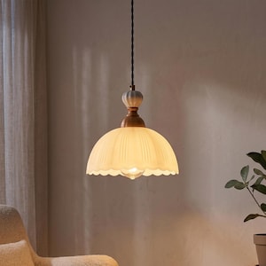 Peut inclure: Une suspension lumineuse dotée d'un abat-jour en verre dépoli festonné de couleur crème. Le luminaire comprend un col doré métallique et une petite pièce décorative en céramique nervurée au sommet, suspendue par un cordon torsadé noir.