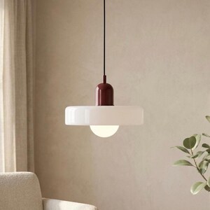 Peut inclure: Un luminaire suspendu moderne avec un abat-jour blanc plat et une source de lumière sphérique. La partie supérieure est de couleur rouge foncé et la lumière est suspendue à un cordon noir. Le fond est beige neutre.