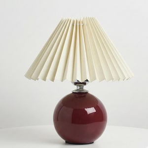 Pode incluir: Candeeiro de mesa bordô com abajur plissado creme. A lâmpada tem uma base esférica e um pescoço cromado. O abajur tem um design em forma de leque. Luminária decorativa.