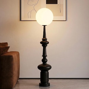 Puede incluir: Una lámpara de pie negra con una pantalla esférica de vidrio blanco. La lámpara tiene una base decorativa de madera torneada y un soporte vertical esbelto. La lámpara es una adición elegante a cualquier sala de estar o dormitorio.