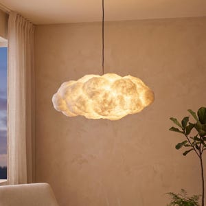 May include: A cloud-shaped pendant light fixture. The light is white and has a fluffy, textured appearance, resembling a cloud. It hangs from a black cord against a neutral-toned wall. The fixture is illuminated, casting a warm glow.