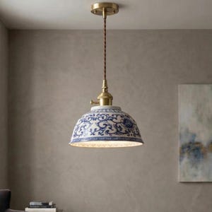 Może przedstawiać: Lampa wisząca z ceramicznym kloszem z niebiesko-białym kwiatowym wzorem. Lampa ma mosiężną osłonę i oprawę z brązowym skręconym przewodem. Klosz ma średnicę około 20 cm.