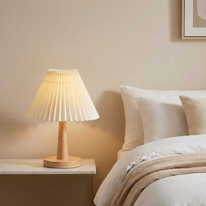 May include: A cream-colored pleated lampshade sits atop a wooden base and stem. The lamp is on a light-colored nightstand next to a bed with white linens and a tan blanket. The lamp provides a warm, inviting glow.