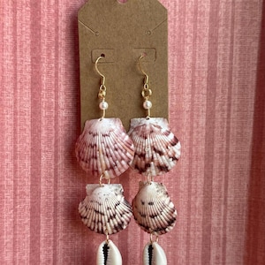 Puede incluir: Pendientes de concha hechos a mano con ganchos y detalles dorados. Los pendientes presentan dos juegos de conchas en tonos rosa, marrón y blanco, con una pequeña concha de cauri blanca colgando de la parte inferior de cada pendiente.