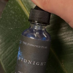 May include: A small clear glass dropper bottle filled with dark blue liquid. The black label features the text: THE POISONED PEARL, MIDNIGHT, DREAM WORK, INTUITION, EMBODIMENT OF THE WATER ELEMENT, EMOTIONAL DEPTH. The bottle has a black ribbed cap.