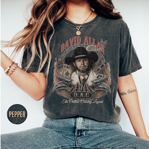 May include: Dark gray t-shirt featuring a graphic of David Allan Coe in a cowboy hat, with guns and playing cards. The text reads "DAVID ALLAN COE" and "D.A.C. The Outlaw Country Legend".