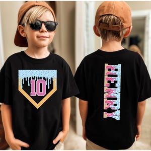 May include: Black t-shirt with a baseball home plate design featuring a light blue frosted top with sprinkles and dripping icing. The number 10 is in the center. The back of the shirt has the name "HENRY" in a vertical design.