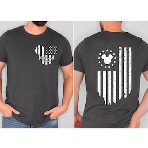 May include: Dark gray t-shirt featuring a Mickey Mouse-themed design. The front displays a Mickey head with stripes and stars, and the text "250 years of Freedom." The back showcases a distressed American flag design with a Mickey head and stars.