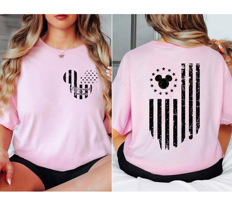 May include: Light pink t-shirt featuring a Mickey Mouse-shaped design with an American flag theme. The front has a Mickey head with stripes and stars, and the text "250 years of Freedom". The back displays a distressed American flag design.