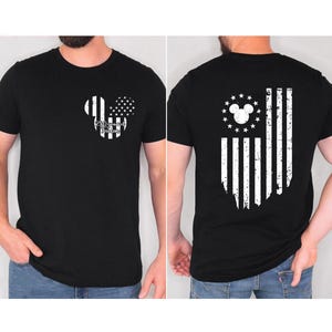 May include: Black t-shirt featuring a distressed American flag design with a Mickey Mouse silhouette. The front displays a Mickey head with flag elements and text. The back shows a flag design with a Mickey head and stars.