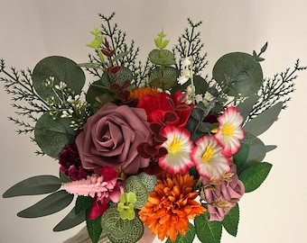 May include: A floral bouquet featuring a large mauve rose, a vibrant orange chrysanthemum, and white flowers with pink-tipped petals. The arrangement includes various green eucalyptus leaves, dark red accents, and feathery pink stems, creating a dense, colorful display of artificial botanical elements.