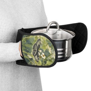 May include: Black quilted oven mitts with a butterfly design. The mitts feature a black and white butterfly on a green and white floral background. A stainless steel pot with a glass lid is held by the mitts.