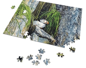 May include: A partially assembled jigsaw puzzle featuring a detailed illustration of a puffin bird perched on a rock. The puzzle showcases the bird with its distinctive orange beak, set against a backdrop of green grass and a rocky cliff. Several puzzle pieces are scattered around.