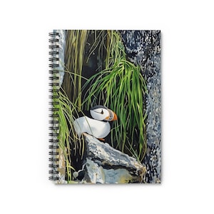 May include: Spiral-bound notebook featuring a watercolor painting of a puffin bird. The bird, with its white, black, and orange beak, is perched on a rock amidst green grass and a dark background. The notebook's cover showcases the artwork.
