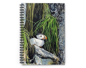 May include: Spiral-bound notebook featuring a watercolor painting of a puffin bird. The bird, with its white, black, and orange beak, is perched on a rock amidst green grass and a dark background. The notebook's cover showcases the artwork.