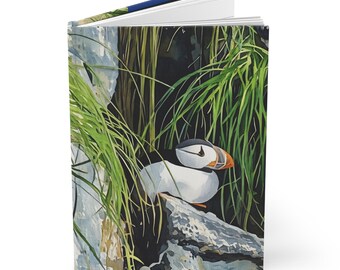 May include: Hardcover journal featuring a watercolor illustration of a puffin bird resting on a rock, surrounded by green grass. The journal has a white interior and a blue spine. The cover art showcases the bird's black, white, and orange beak.