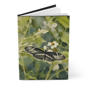 May include: Hardcover journal featuring a watercolor painting of a butterfly with black and white stripes resting on white flowers. The background has green foliage and a watercolor wash.