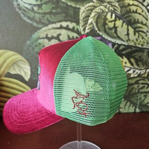 Op de afbeelding: Een baseballcap met een roze klep en een rode bovenkant. De achterkant van de cap is groen mesh. De cap heeft een rood geborduurd ontwerp aan de zijkant. De cap wordt op een transparante standaard getoond.