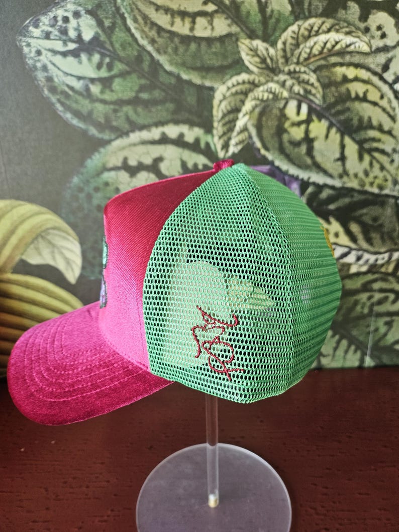 Op de afbeelding: Een baseballcap met een roze klep en een rode bovenkant. De achterkant van de cap is groen mesh. De cap heeft een rood geborduurd ontwerp aan de zijkant. De cap wordt op een transparante standaard getoond.