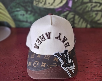 Op de afbeelding: Crème baseballpet met "BAY AREA" in bruine letters. De klep is bruin met een herhalend patroon. Een bruin en witte patch met een druipend ontwerp staat aan de zijkant.