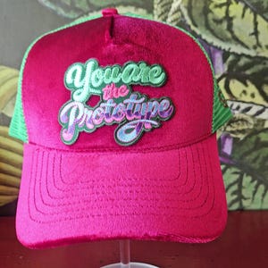 May include: A magenta trucker hat with a green mesh back and a curved brim. The hat features the embroidered text "You are the Prototype" in green, purple, and blue lettering. The hat is displayed on a clear stand.