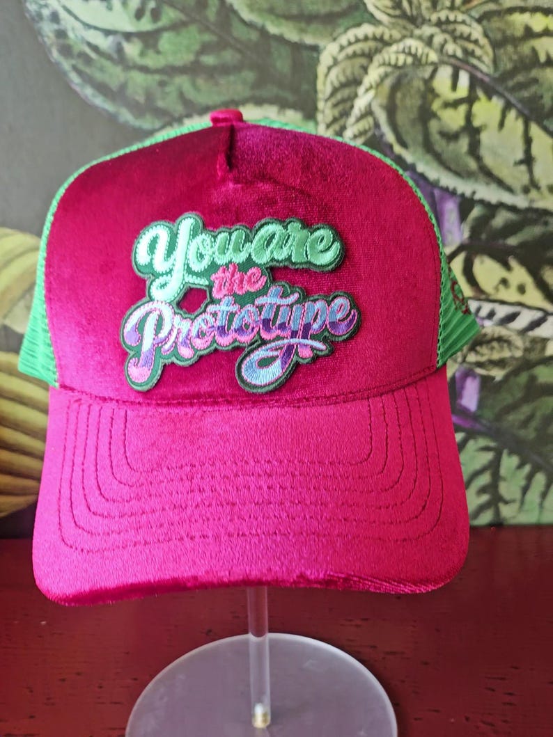 Op de afbeelding: Een magenta truckerpet met een groene mesh achterkant en een gebogen klep. De pet heeft de geborduurde tekst "You are the Prototype" in groene, paarse en blauwe letters. De pet wordt op een transparante standaard getoond.
