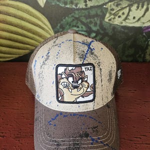 May include: Brown and tan trucker cap with a Taz cartoon patch. The cap has a mesh back, a curved peak, and blue paint splatter details. The Taz patch is square and features the cartoon character.