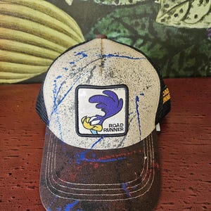 Puede incluir: Gorra de camionero con visera marrón y corona beige, decorada con salpicaduras de pintura azul y roja. La gorra presenta un parche con el personaje de dibujos animados Road Runner y las palabras "Road Runner". La parte trasera es de malla negra.