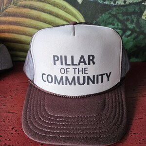 May include: A brown and white trucker hat with the text "PILLAR OF THE COMMUNITY" in black. The hat has a brown brim and a white front panel with the text. The sides and back are gray mesh.