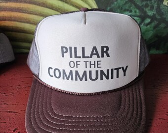 Op de afbeelding: Bruine en witte truckerpet met de tekst "PILLAR OF THE COMMUNITY" in zwart. De pet heeft een bruine klep en een wit voorpaneel met de tekst. De zijkanten en achterkant zijn van grijs mesh.
