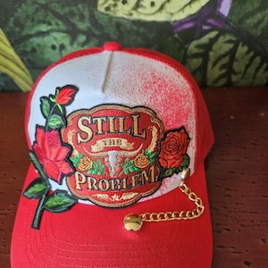 May include: Red and white trucker cap with a red peak and mesh back. The front has an embroidered patch with the words "Still The Problem" and rose details. A gold chain hangs from the side.