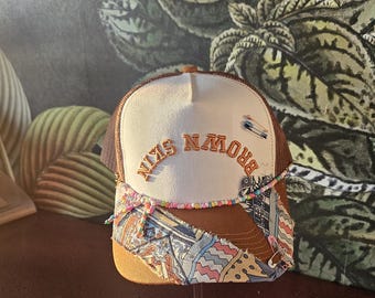 Op de afbeelding: Truckerpet met een wit voorpaneel en een bruine mesh achterkant. De pet heeft de tekst "BROWN SKIN" in geborduurde oranje letters. De rand is een patchwork van stoffen met patronen. Een kralenketting en veiligheidsspelden sieren de pet.