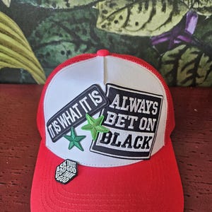 May include: Red and white trucker hat with a curved brim. The hat features patches with the phrases "IT IS WHAT IT IS" and "ALWAYS BET ON BLACK". Includes a pin that says "ASK GOD TRUST GOD THANK GOD".
