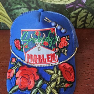 Könnte beinhalten: Blaue Trucker-Cap mit Mesh-Rückseite, bestickt mit roten Rosen und dem Text "Somebody's Problem". Die Cap hat einen Strassbesatz und eine dekorative Nadel mit blauen Perlen.