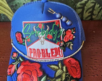 Może przedstawiać: Niebieska czapka typu trucker z siatkowym tyłem, z haftowanymi czerwonymi różami i napisem "Somebody's Problem". Czapka jest ozdobiona obszyciem z dżetów i ozdobną szpilką z niebieskimi koralikami.