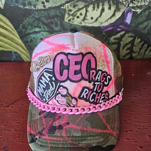 May include: A camouflage trucker hat with pink accents and patches. The hat features the text "CEO", "Rags to Riches", and "Mama Tried". A pink chain adorns the brim, and the crown has a pink and white splatter design.